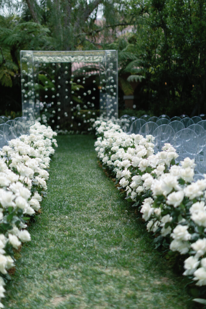 Outdoor wedding aisle lined with white roses, leading to a transparent altar under a tree canopy. Transparent chairs are arranged on either side.