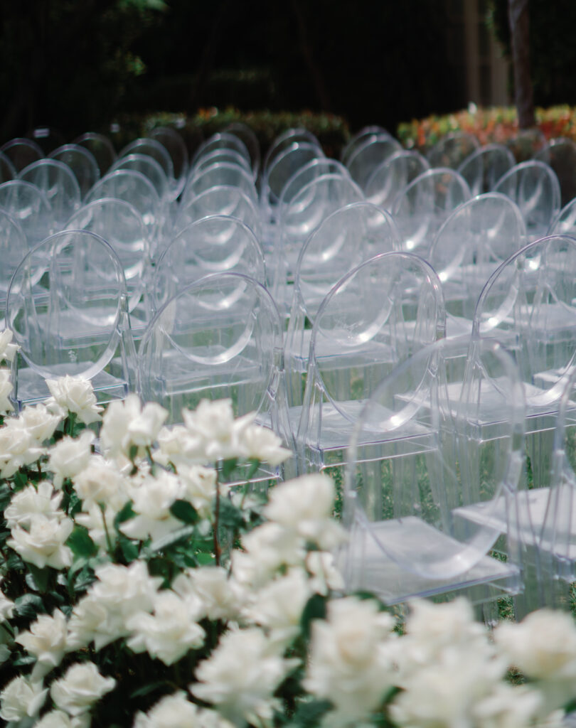 Rows of clear chairs set up outside with white flowers in the foreground.