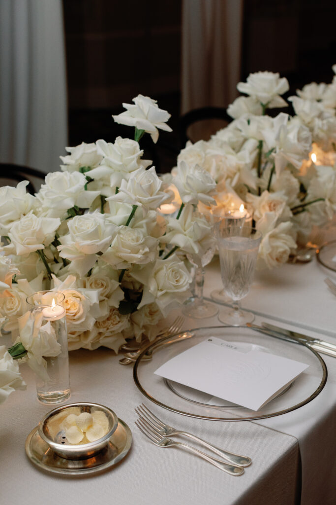 Elegant table setting with white roses, candles, crystal glassware, a silver plate with butter, and a white napkin on a beige tablecloth.