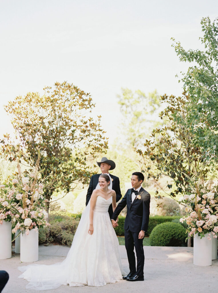 A couple stands together in wedding attire, holding hands outdoors, with a person officiating behind them. Bushes and floral arrangements surround the scene.