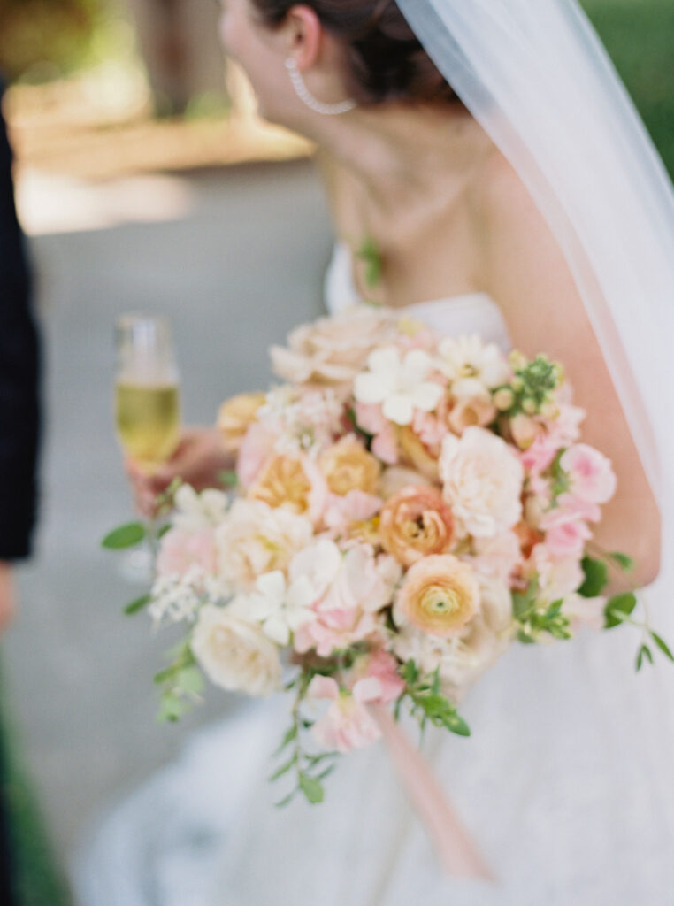 Bride holding a bouquet of pink, peach, and white flowers, and a glass of champagne.