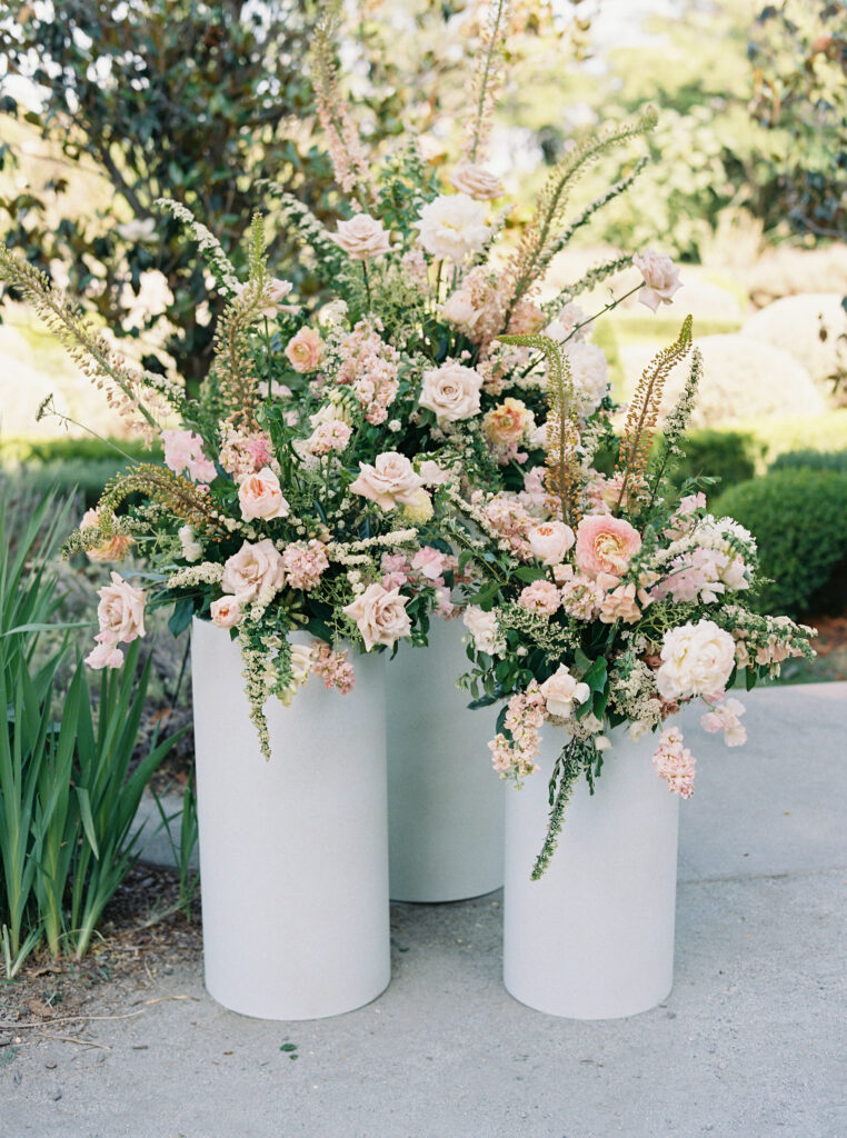 Three white cylindrical vases hold arrangements of pink and white flowers with greenery, set outdoors on a stone surface.