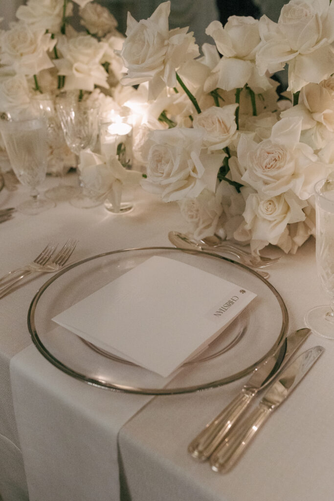 Elegant table setting with a transparent plate, silver cutlery, and a folded menu card. White roses and candles decorate the table.