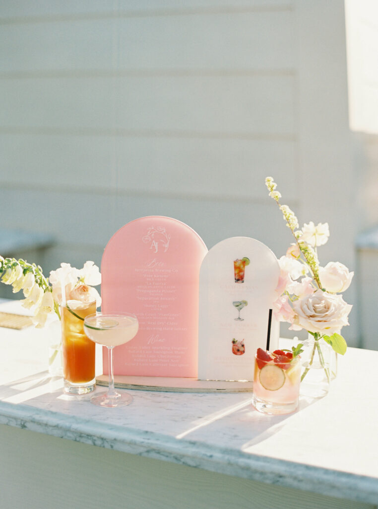 A menu with wedding drinks is displayed on a marble counter, featuring three cocktails in glasses with garnishes, surrounded by pink and white flowers.