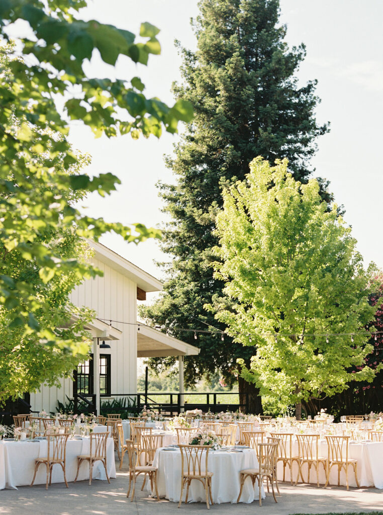 Outdoor event setup with round tables covered in white tablecloths and wooden chairs beneath trees next to a white building.