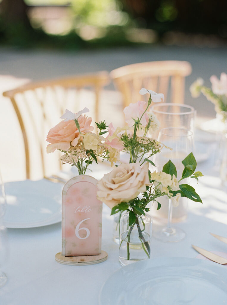 Table setting with a floral centerpiece featuring pale orange and cream roses, accompanied by a table number 6 sign, set on a white tablecloth with glassware and cutlery.