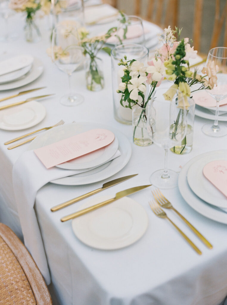 A table set for a formal meal with white plates, gold cutlery, pink menus, and small flower arrangements in glass vases.