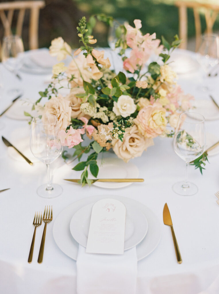 A round table with a white tablecloth is set with gold cutlery, a white plate, wine glasses, and a floral centerpiece of pastel flowers. A menu is placed on the plate.