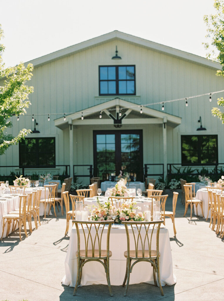 Outdoor wedding reception setup in front of a white barn. Tables are adorned with white tablecloths and floral centerpieces. String lights hang above, and wooden chairs surround the tables.