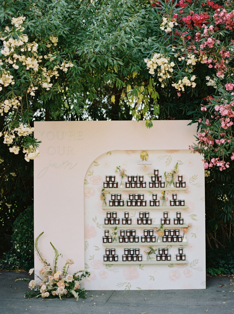 A decorative display with small bottles arranged on shelves against a floral backdrop, surrounded by blooming flowers and greenery. The words "You’re Our Jam" are written on the panel.