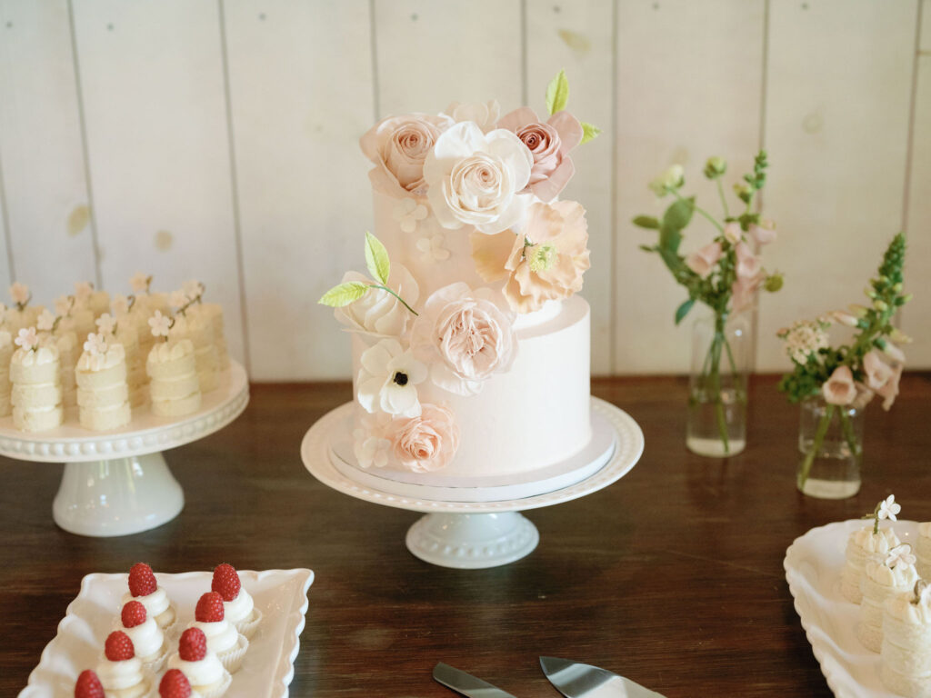 A two-tiered white cake with pink and white floral decorations on a cake stand, surrounded by desserts and flowers.