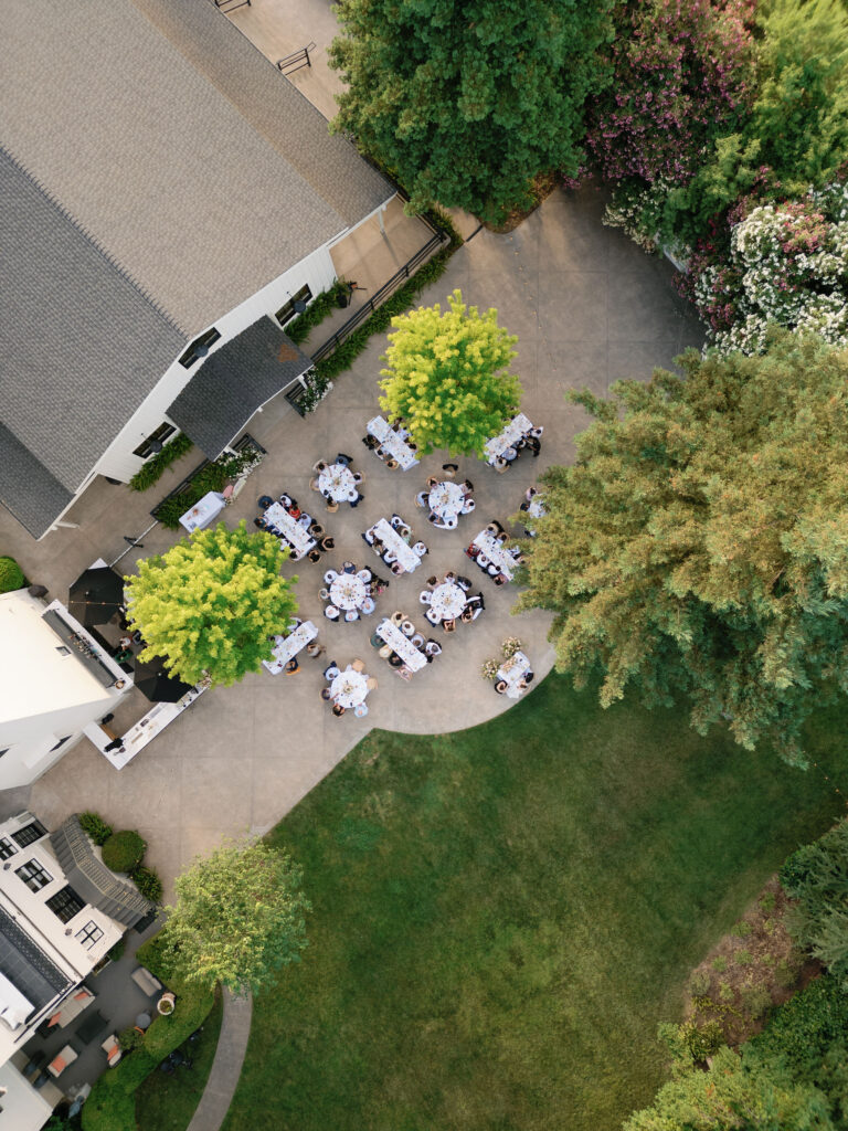 Aerial view of an outdoor wedding reception with several tables set for guests, situated on a concrete patio surrounded by green trees and landscaped lawn.