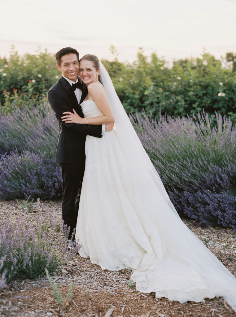 A bride and groom in wedding attire pose smiling in a garden with purple flowers and greenery in the background.