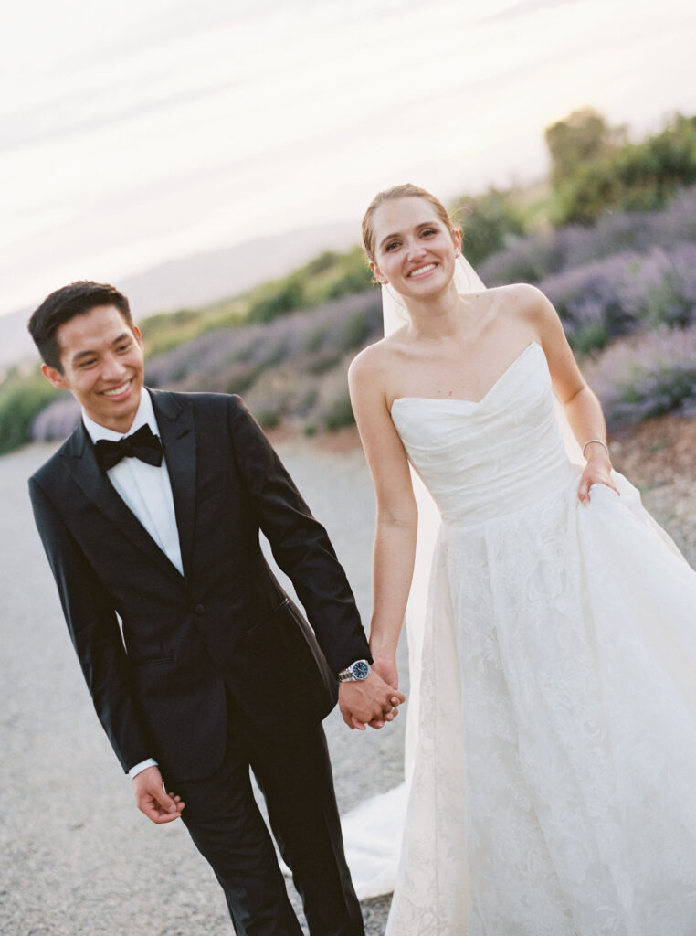 A bride in a white dress and a groom in a black suit hold hands while walking on a gravel path, with greenery in the background.