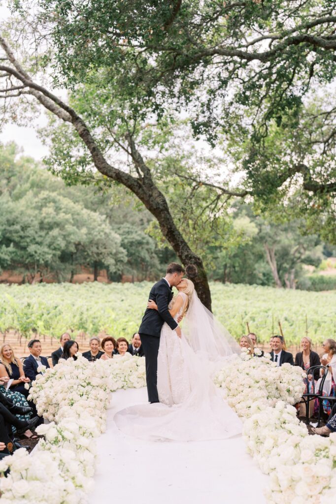 A bride and groom kiss at an outdoor wedding, surrounded by white flowers and guests, with a vineyard and trees in the background.