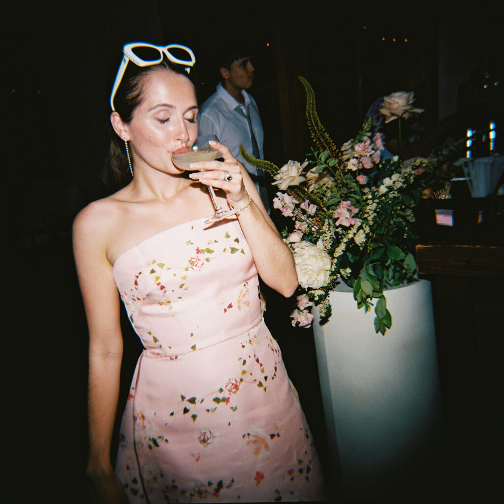 Woman in a floral dress sips a drink near a flower arrangement.