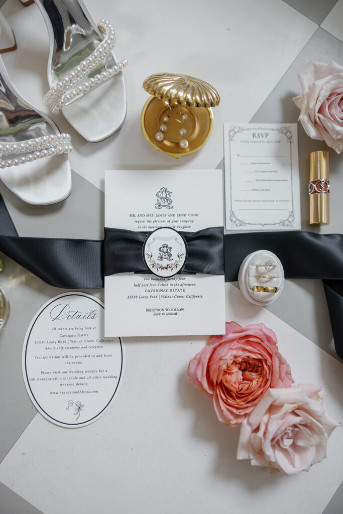 Wedding invitation set with envelopes, RSVP card, pearl-accented shoes, rose flowers, jewelry box with rings, lipstick, and pearl detail.