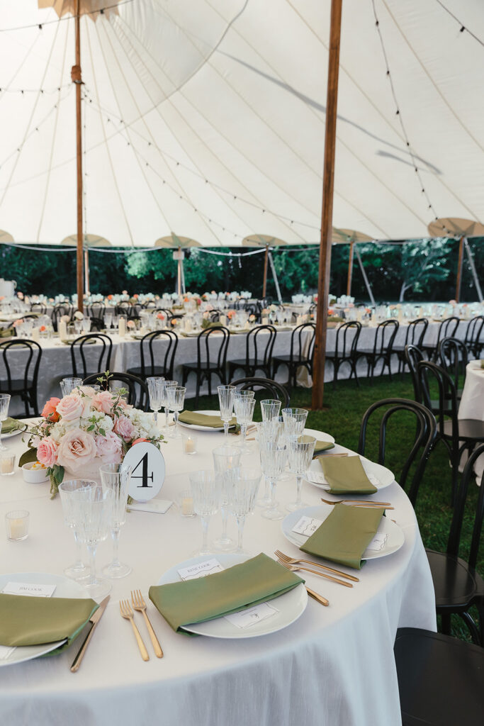 Wedding reception tent with round tables set with green napkins, glassware, and floral centerpieces under string lights.