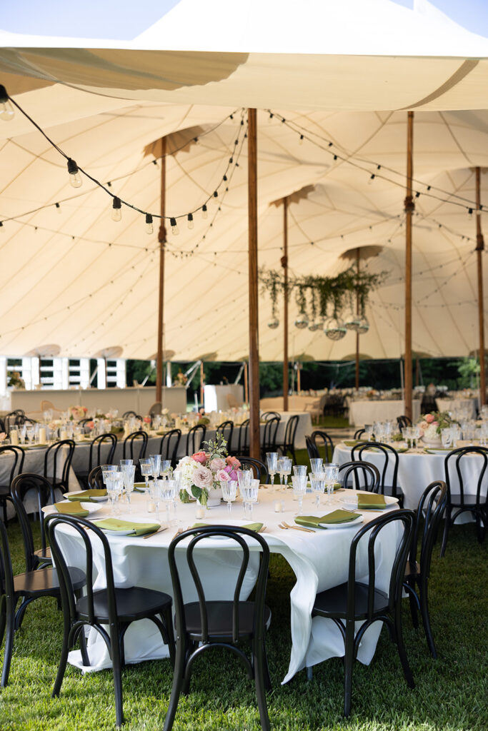 Outdoor event setup under a large tent with round tables, black chairs, white tablecloths, floral centerpieces, and string lights above.