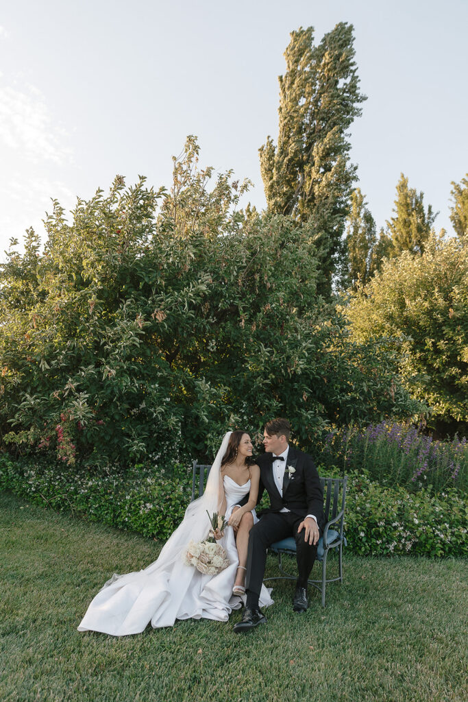 A bride and groom sit on a bench in a garden. The bride wears a white dress and holds a bouquet, while the groom wears a black suit. They are looking at each other.