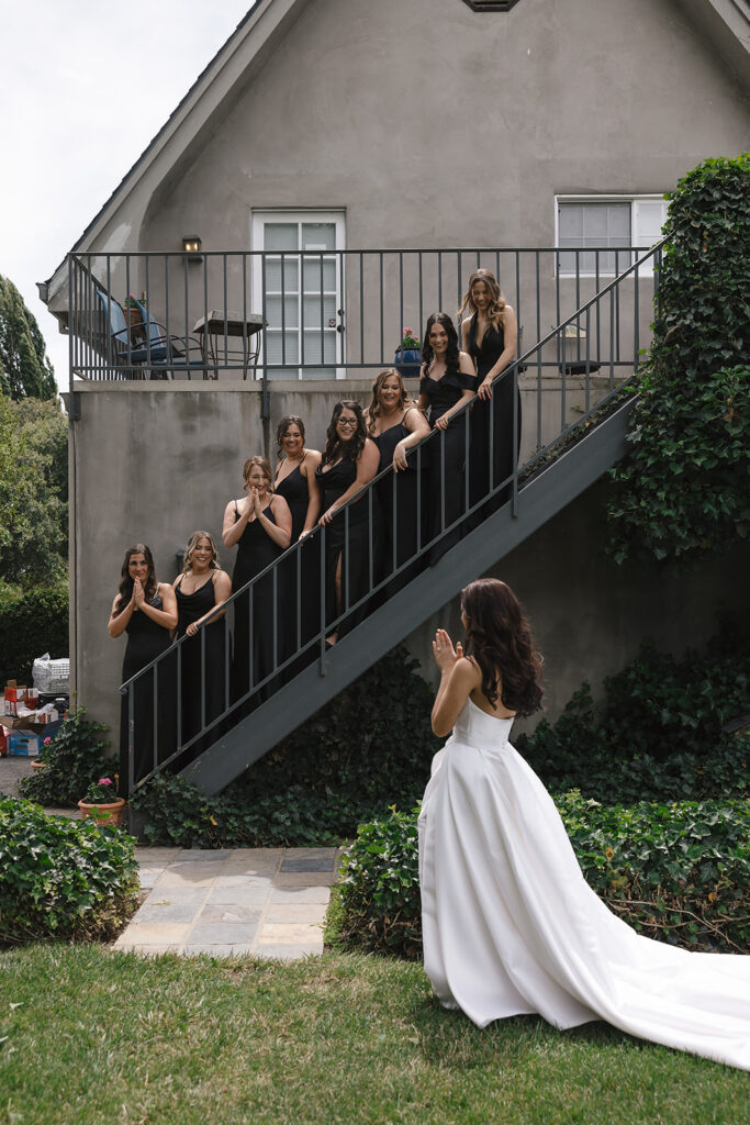A bride in a white dress stands at the bottom of outdoor stairs, while seven bridesmaids in black dresses pose along the steps, looking towards her.