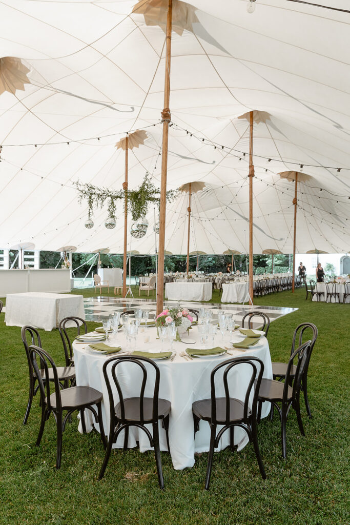 An outdoor event tent with a round table set for dinner, surrounded by dark wooden chairs on a grassy area. The tent is decorated with plants and string lights.