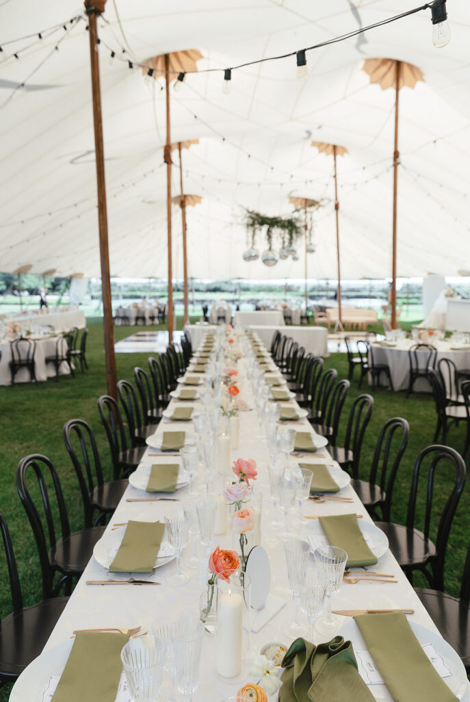 Long table set for an event under a white tent, featuring green napkins, glassware, and floral centerpieces with chairs on either side.