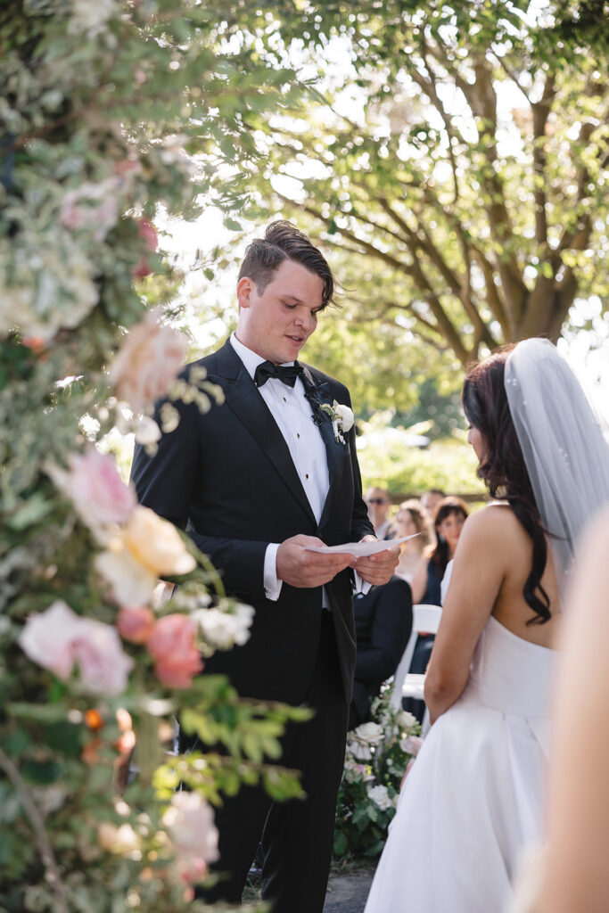 A groom in a tuxedo reads vows to the bride in a white dress during an outdoor wedding ceremony under trees.