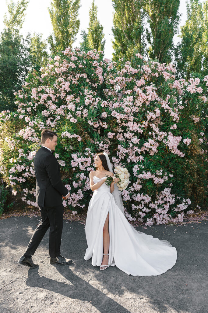 A bride in a white dress and a groom in a black suit stand in front of a large bush with pink flowers.
