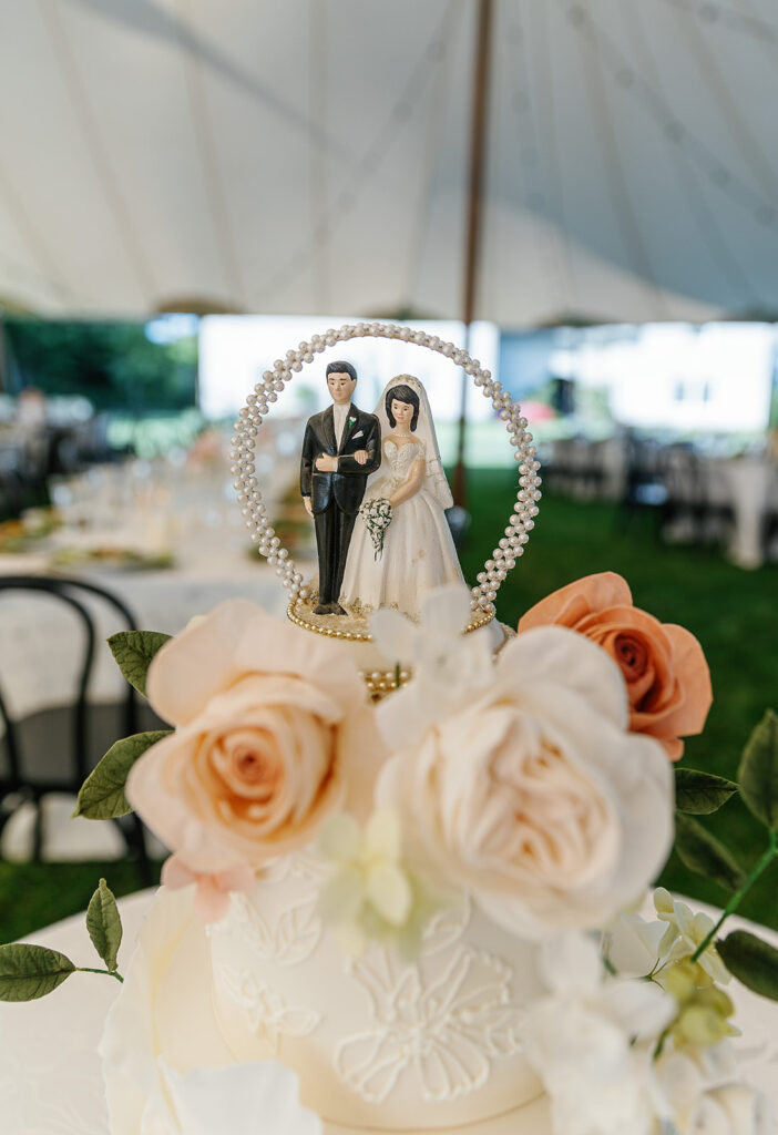 A wedding cake topped with a bride and groom figurine is adorned with large cream and peach roses, under a white tent at an outdoor event.
