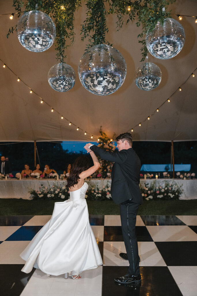 A couple dances on a checkered floor under hanging disco balls in a decorated tent.