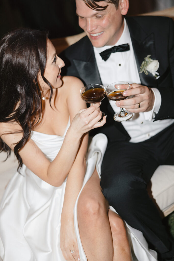 A couple in formal attire toasts with champagne glasses, seated closely.