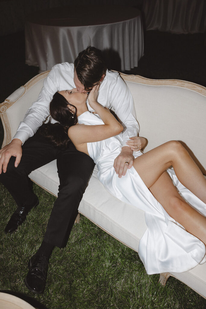 A couple in white formal attire kisses on a cream-colored sofa placed on a grassy area.
