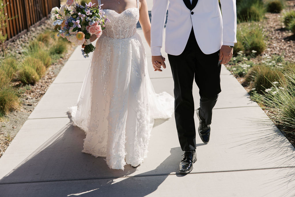 A bride in a white dress and a groom in a white jacket and black pants hold hands while walking on a path, with the bride carrying a bouquet of flowers.