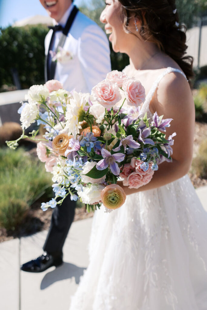 A bride and groom walk outdoors, smiling. The bride holds a colorful bouquet with pink, blue, and white flowers.
