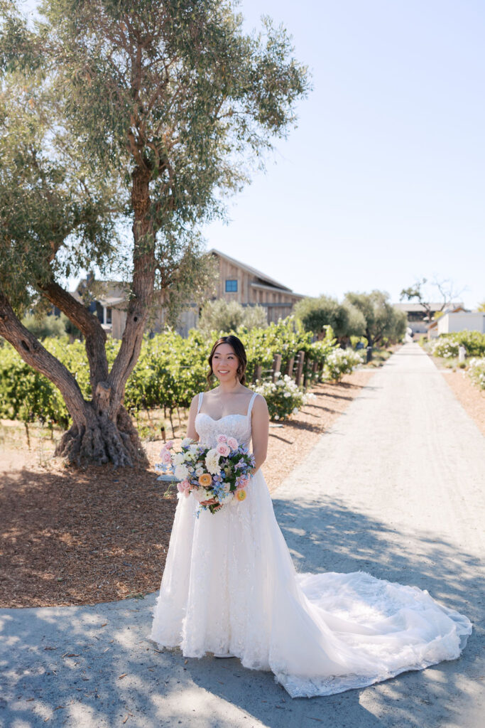 A bride in a white gown stands on a gravel path holding a bouquet, with a vineyard and house in the background.