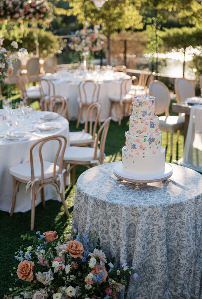 A multi-tiered floral cake on a decorated table in an outdoor setting, surrounded by elegantly set tables and chairs with floral arrangements.