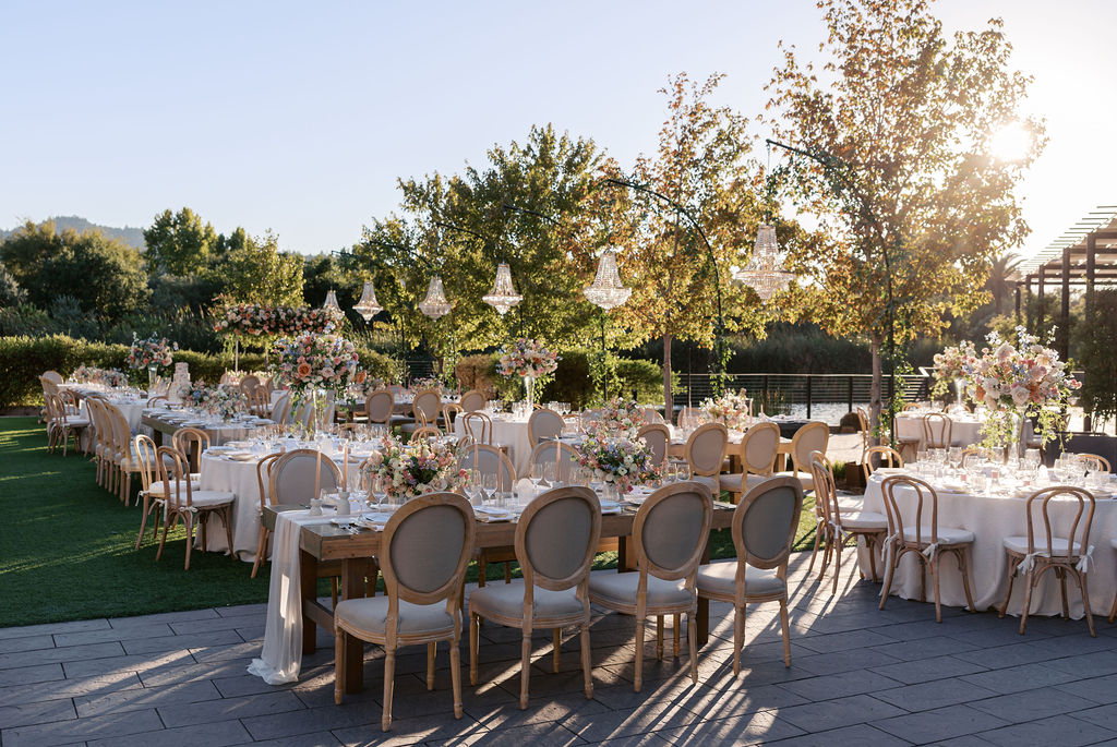 Elegant outdoor wedding setup with long tables, floral centerpieces, wooden chairs, and chandeliers, under a canopy of trees with sunlight filtering through.
