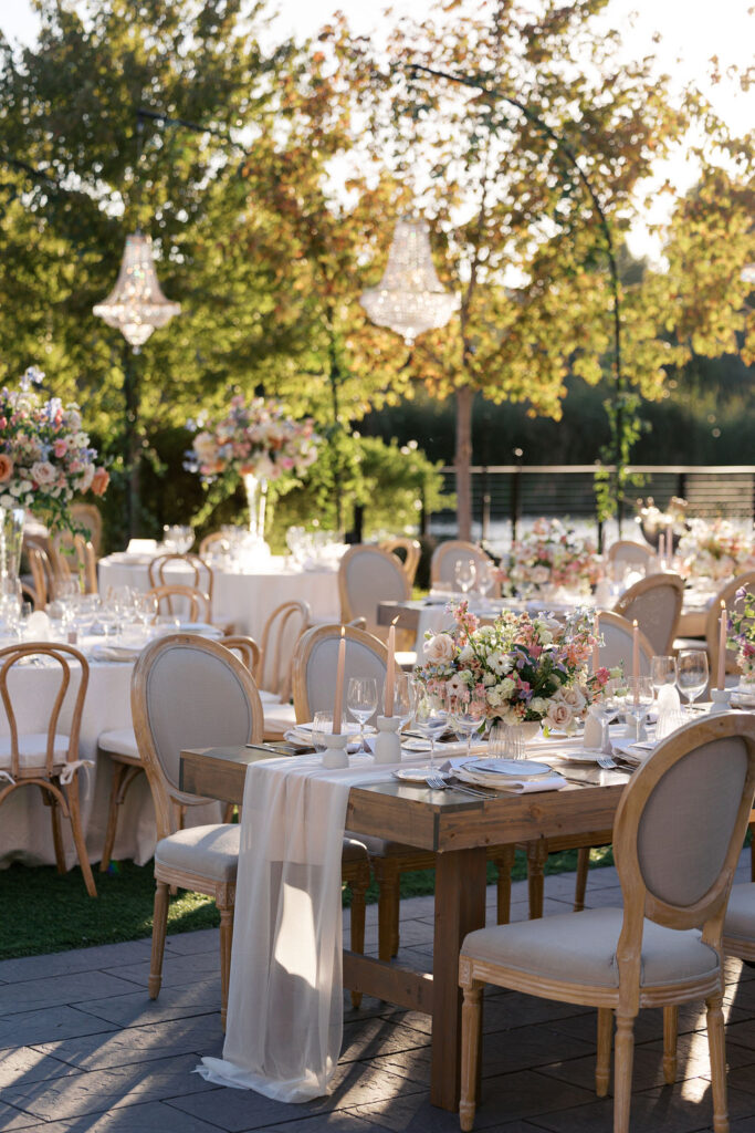 An elegant outdoor event setup with decorated tables, floral centerpieces, and chandelier lights under a canopy of trees.
