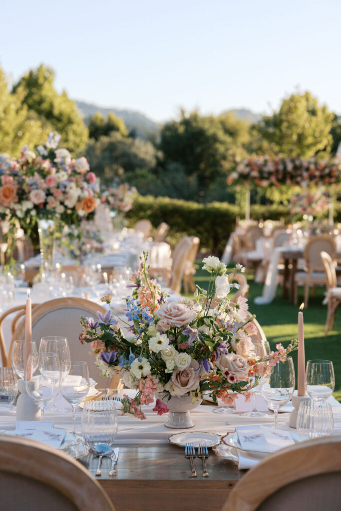 An elegant outdoor dining setup with floral centerpieces, glassware, and neutral-colored candles on tables surrounded by chairs. Lush greenery and trees are visible in the background.
