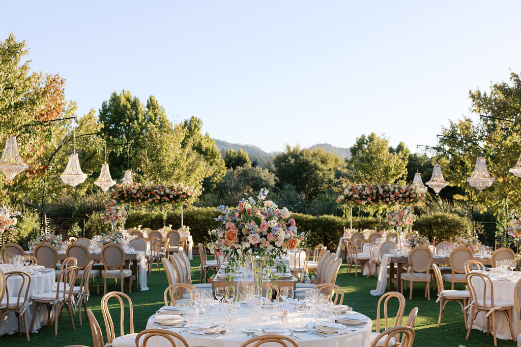 Outdoor wedding reception with round tables, elegant floral centerpieces, and chandeliers, set on green lawn surrounded by trees.