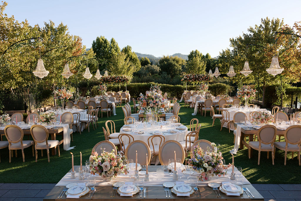 An elegant outdoor wedding reception with tables set for dining, adorned with floral centerpieces and chandeliers. Surrounded by trees on a sunny day.