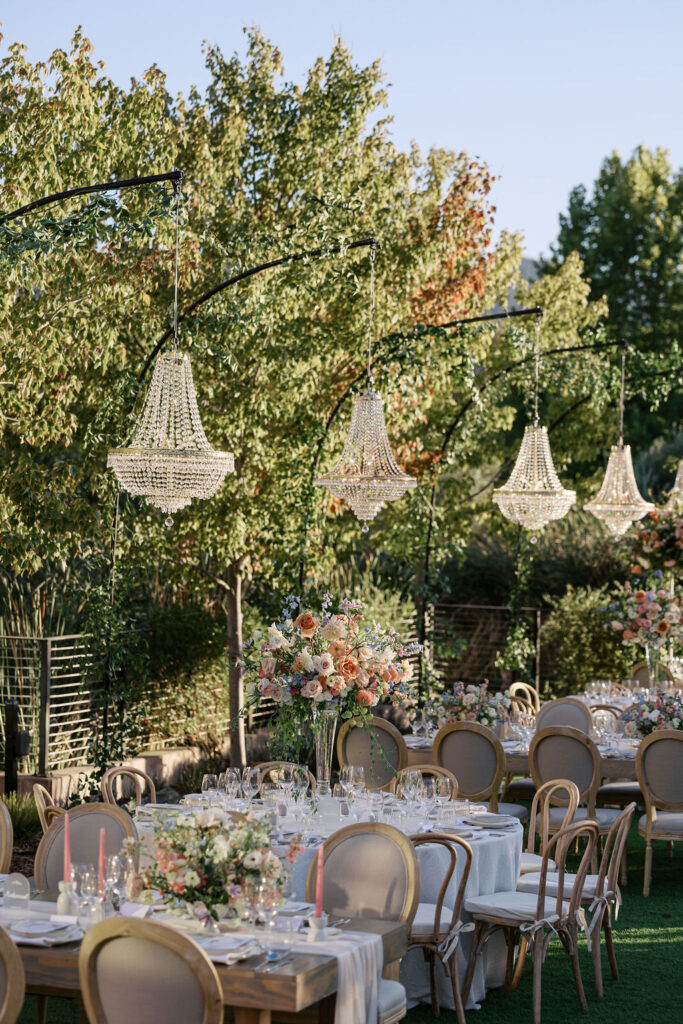 Outdoor wedding reception with tables set for dining, featuring floral arrangements and chandeliers hanging above. Trees and a clear sky form the backdrop.
