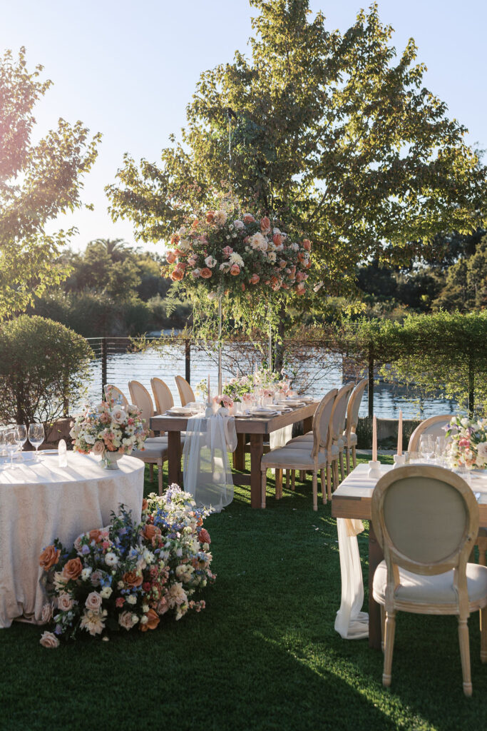 Outdoor wedding setup with decorated tables and chairs on a lawn by a lake. Floral arrangements adorn the tables, and sunlight filters through the trees.