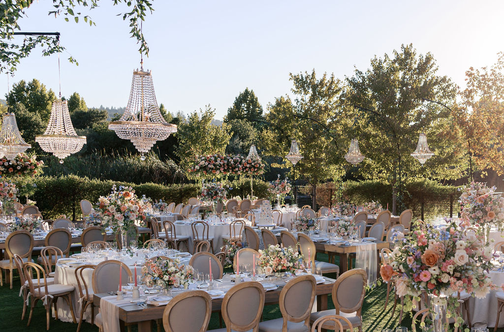 Outdoor wedding reception with elegantly set tables, floral centerpieces, and chandeliers hanging from above. Trees and greenery surround the area.