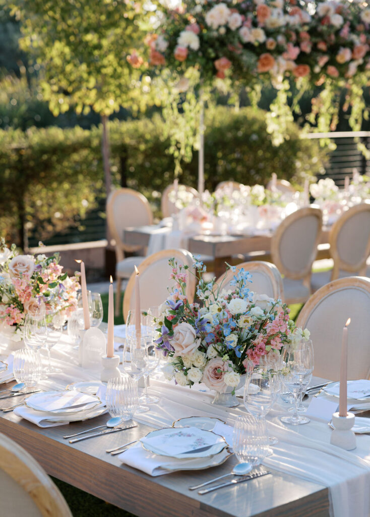 Elegant outdoor dining table setup with floral centerpieces, candles, and place settings. Surrounded by greenery and softly lit by natural sunlight.