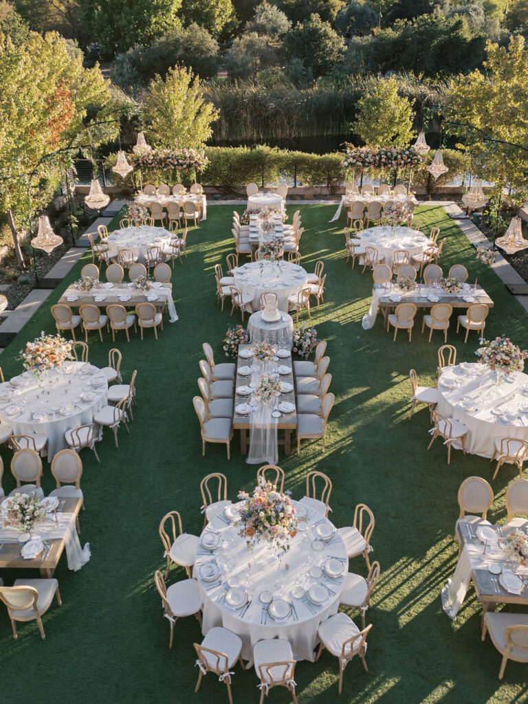 Outdoor wedding reception setup with round and rectangular tables covered in white tablecloths, surrounded by chairs. Tables are decorated with floral centerpieces, set on a grassy area.
