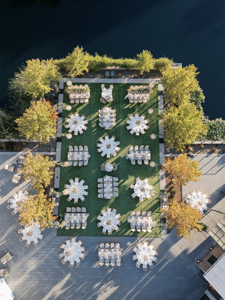 Aerial view of an outdoor event setup with round tables and chairs on a green lawn, surrounded by trees and near a body of water.