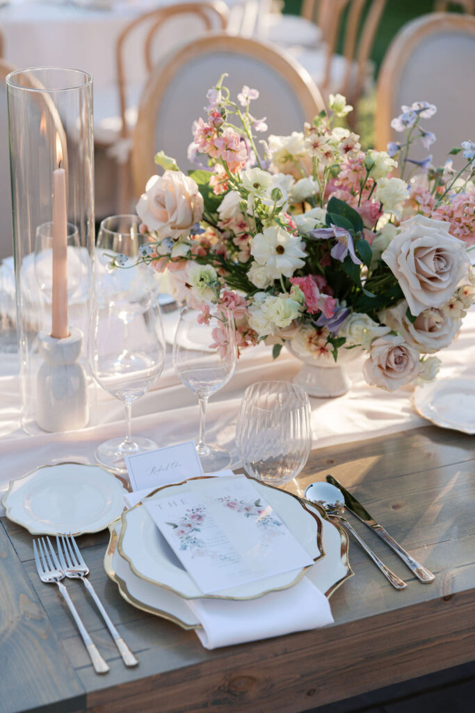 Elegant table setting with floral centerpiece, white plates, crystal glassware, and gold cutlery on a wooden table. Tall candle in a glass holder beside the arrangement.
