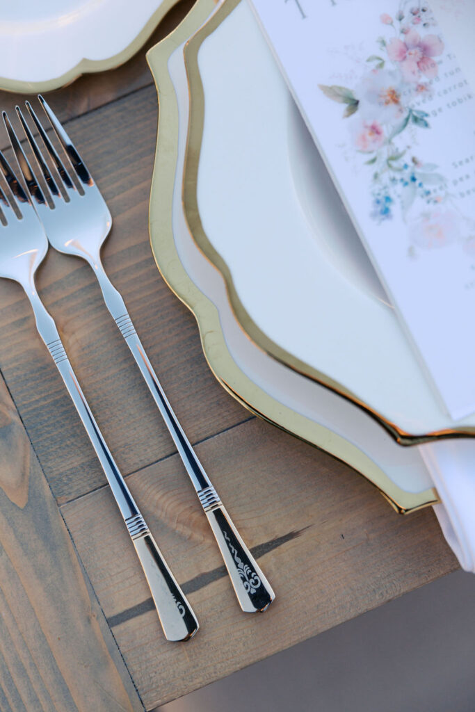 Place setting with two forks, scalloped white plates with gold edges, and a floral menu on a wooden table.
